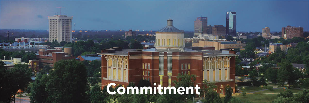A photo overlooking a large building with a red brick design, there is a round center extending higher out of the center of the building. There is text at the bottom of the image that reads "Commitment"