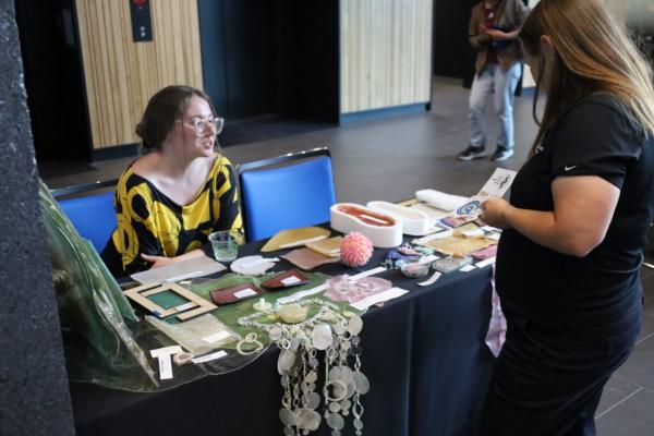 Woman sits at exhibitor table with samples of bioplastics on display