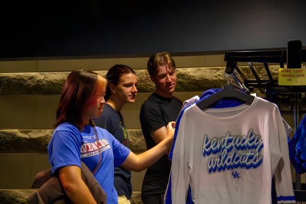 alt="Three students browse a clothing rack, with a white “Kentucky Wildcats” shirt displayed in the foreground."