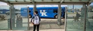 A student waiting in front of the bus stop for a University of Kentucky branded bus. 