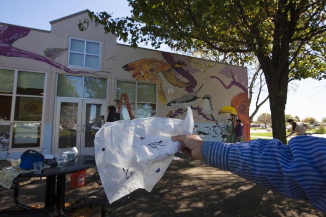 A 1st person POV photo of an arm holding a piece of paper with a bird on it in front of a building where three students work to paint a mural of birds.