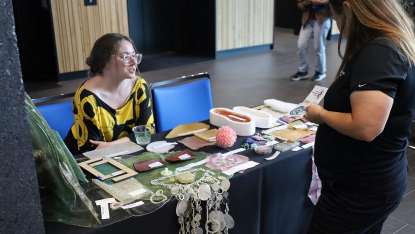 Woman sits at exhibitor table with samples of bioplastics on display